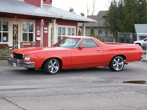 1973 Ford Ranchero - Driver-side 3/4 front view of a bright red 1970s Ford Ranchero coupe-utility parked on pavement in front of a red barn-style building. It r…
