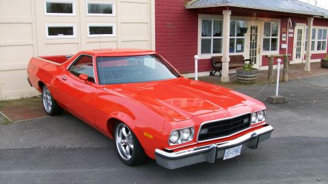1973 Ford Ranchero - Bright red Ford Ranchero car-based pickup viewed from the front three-quarter right, parked on asphalt in front of a red-sided building and…