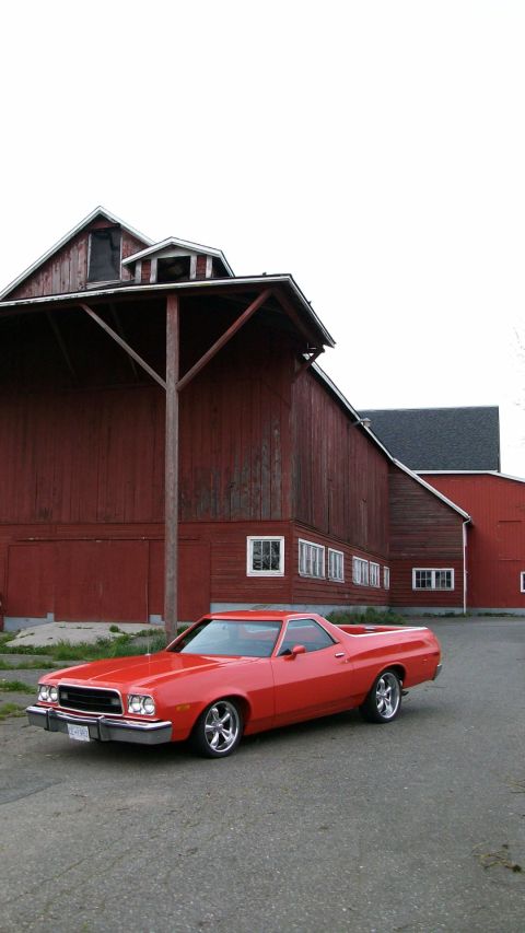 1973 Ford Ranchero - A red 1973 Ford Ranchero is parked on a paved surface in front of a large red barn.