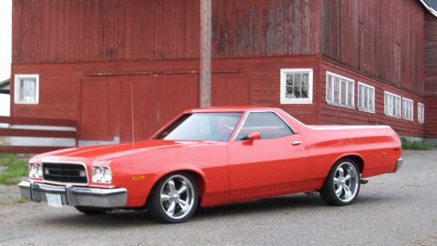 1973 Ford Ranchero - A red 1973-era Ford Ranchero coupe-utility parked on pavement in front of a weathered red barn, shown from the front three-quarter. It has…