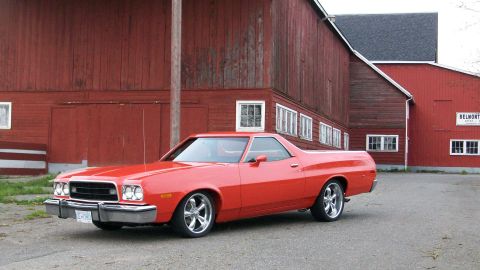 1973 Ford Ranchero - Driver-side front three-quarter view of a lowered orange-red Ford Ranchero coupe-utility on polished five-spoke wheels, parked on gravel-st…