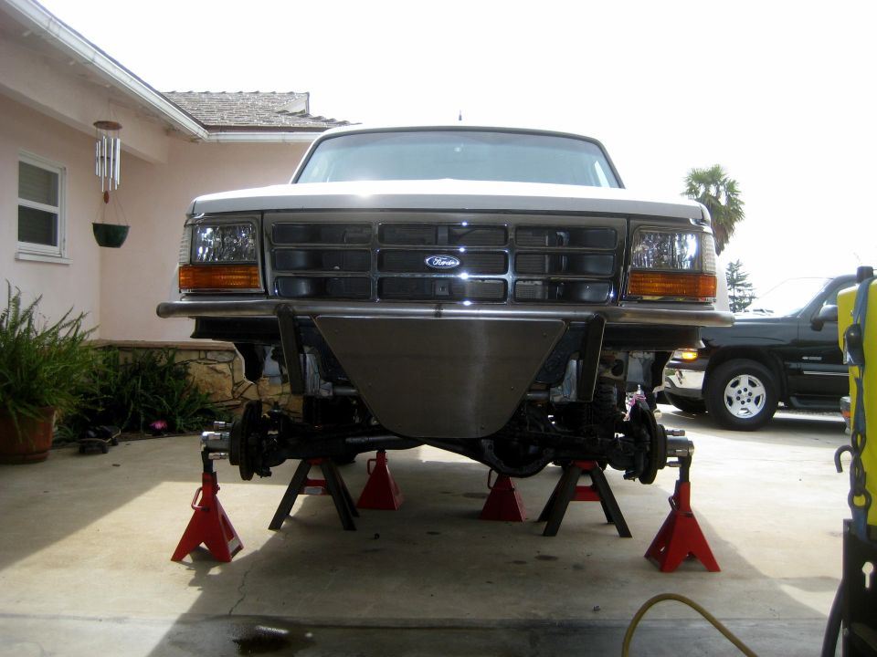 Front of a 1992 Ford truck with a custom bumper, raised on red jack stands. Another dark SUV is parked behind it