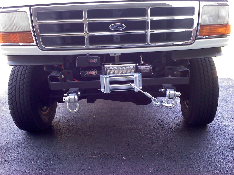 Front of a Ford truck with a chrome grille, Ford emblem, and a winch mounted below the bumper. Tow hooks are visible on either side