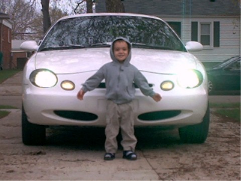 1997 Ford Taurus - A child stands in front of a white 1997 Ford Taurus, its headlights illuminated.