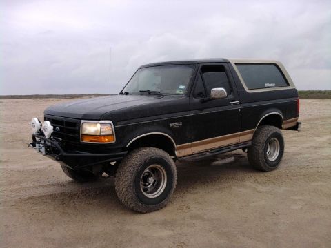 1994 Ford Bronco - This black Ford Bronco has a tan stripe and a custom bumper with auxiliary lights, seen on a sandy terrain.