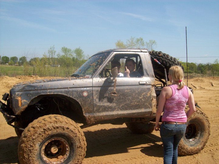 Mud-covered off-road vehicle with large tires, spare tire on back, and two people inside
