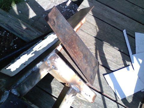1990 Ford Bronco - Close-up of a home-fabricated steel bracket in progress on a weathered wood deck, showing fresh weld beads joining flat bar and gusset plat…
