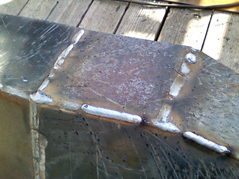 1990 Ford Bronco - Extreme close-up of stitch-welded seams on a fabricated bare-steel plate bracket sitting on weathered deck boards, showing stacked MIG bead…