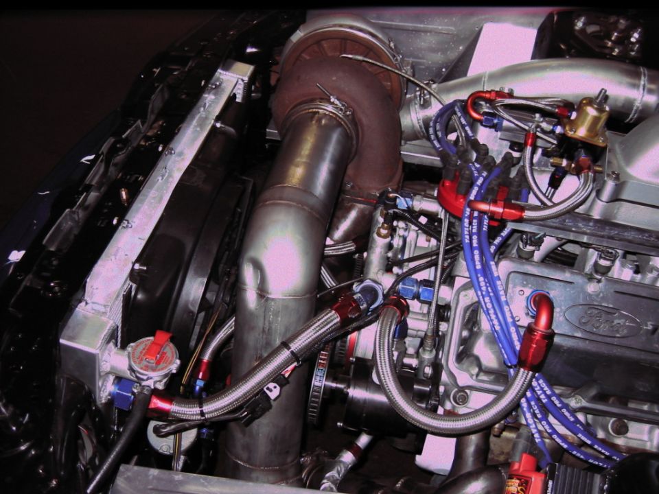 Engine bay with large turbocharger, custom exhaust piping, and braided fuel lines. Ford logo visible on engine block