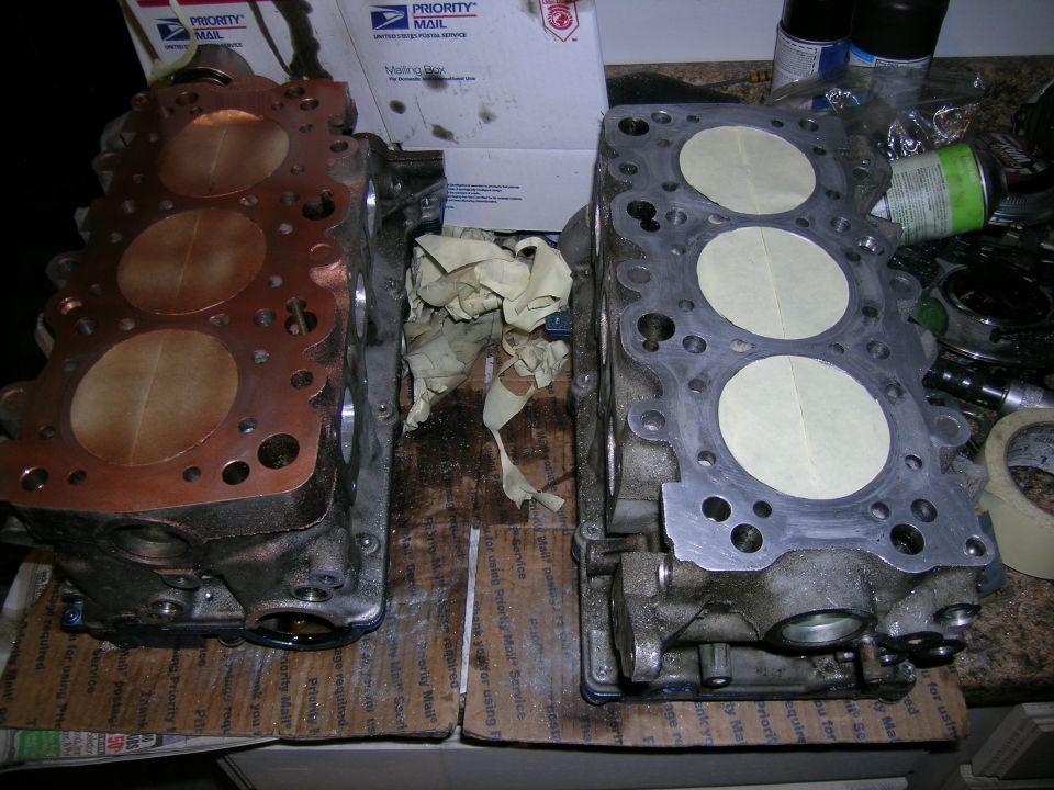 Two engine cylinder heads on a wooden surface, one with copper gasket material, the other with taped-over cylinders