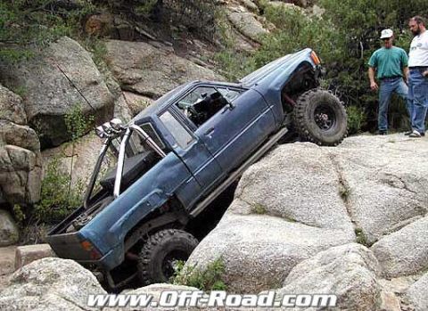 2002 Ford F-350 - A blue off-road truck navigates a challenging rocky terrain, with two onlookers observing the difficult climb.