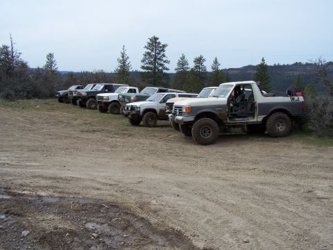 1991 Ford Bronco - A collection of modified trucks and SUVs are parked on a dirt road in a wooded, hilly area.