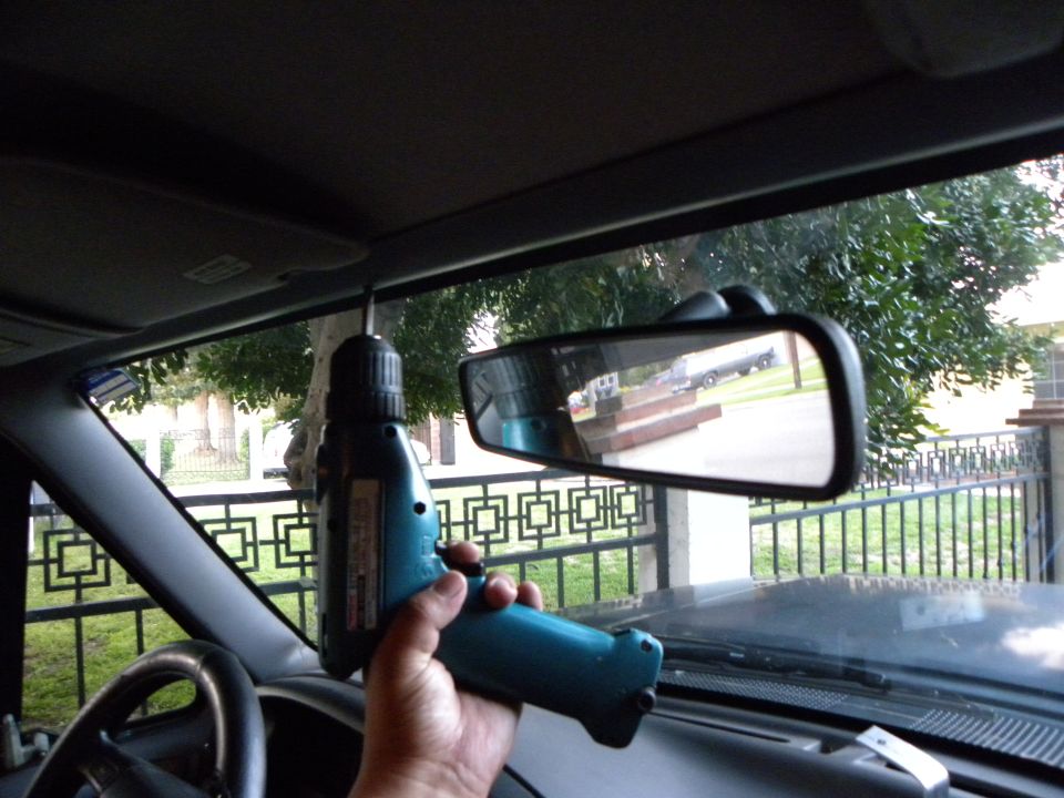 Interior view of a vehicle with a drill being used near the rearview mirror. A hand holds the drill. The mirror reflects a truck