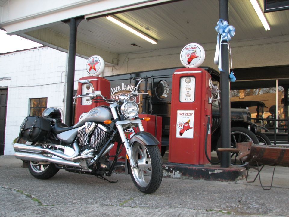 Silver motorcycle parked at vintage gas station with two red gas pumps and a classic car in the background