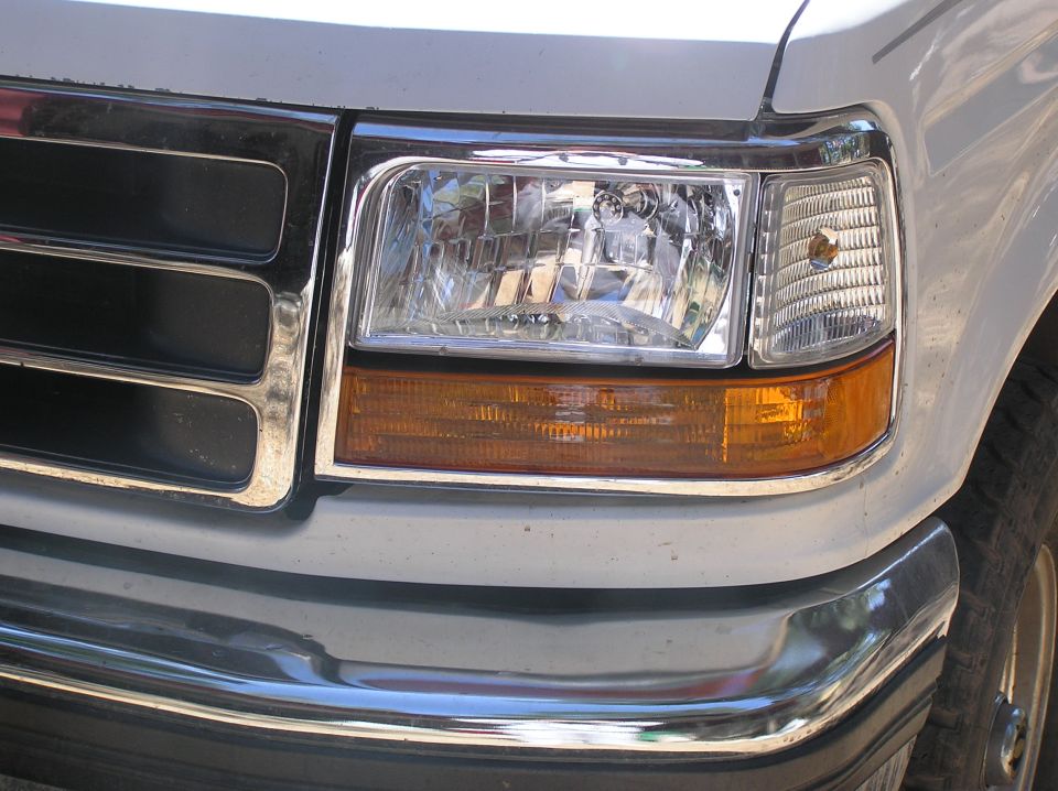 Close-up of a white Ford F-Series truck's front grille and headlight assembly. The grille has horizontal chrome bars