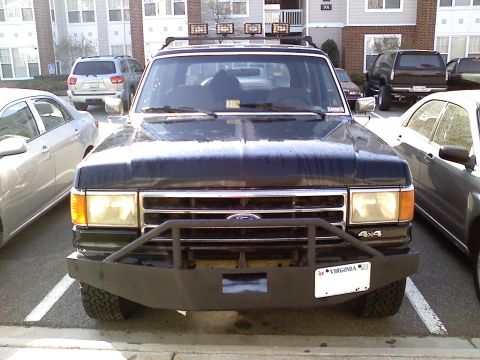 1990 Ford Bronco - Head-on view of a dark blue Ford full-size 4x4 SUV parked between sedans in an apartment complex lot, fitted with a heavy fabricated flat-p…