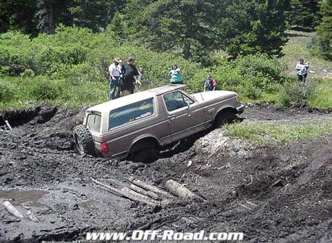 2002 Ford F-350 - A brown Ford Bronco navigates a challenging muddy terrain, with onlookers observing the off-road adventure.