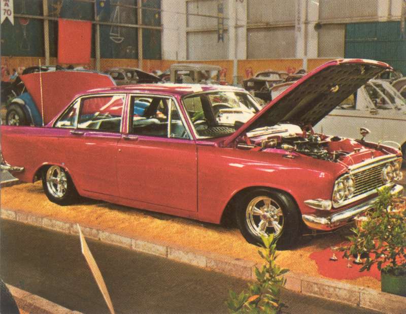 Red customized sedan with hood and trunk open, chrome wheels, parked indoors. Another white vehicle is visible behind it