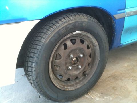 1991 Ford Festiva - Close-up of a blue car's wheel and fender, revealing a rusty steel rim and a NEXEN tire.