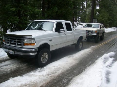 1991 Ford Bronco - A white Ford F-250 pickup truck tows a silver Ford Bronco SUV on a snow-covered road in a forest.