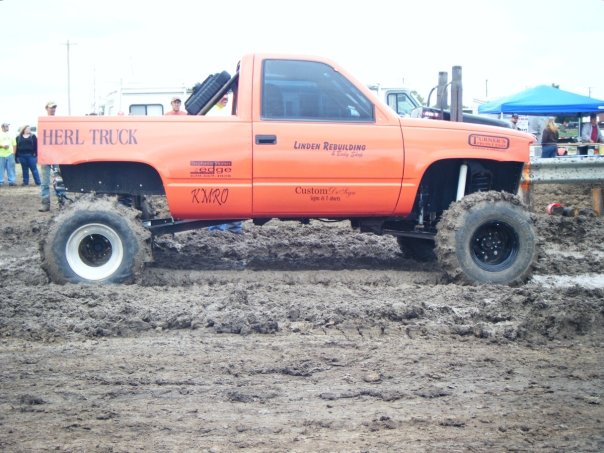 Orange mud bog truck with large tires stuck in mud. Text on side says 'HERL TRUCK' and 'LINDEN REBUILDING'