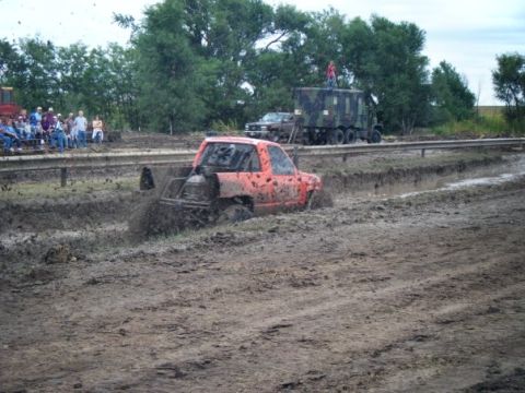 2000 Dodge 1500 - A modified orange pickup truck navigates a muddy track, with spectators and a large military vehicle observing the event.