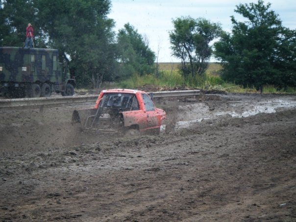 Orange pickup truck driving through a mud pit. A large military-style truck is in the background