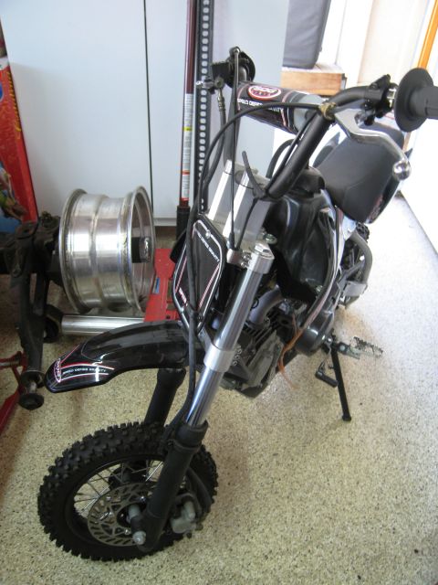 1986 Ford Bronco - A black pit bike is shown from the front, resting on its kickstand with a spare wheel and jack in the background.