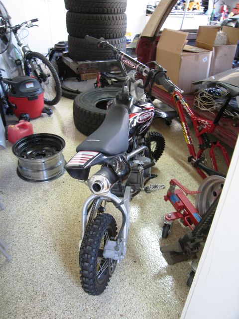 1986 Ford Bronco - A dirt bike sits in a cluttered garage next to spare tires and other equipment.