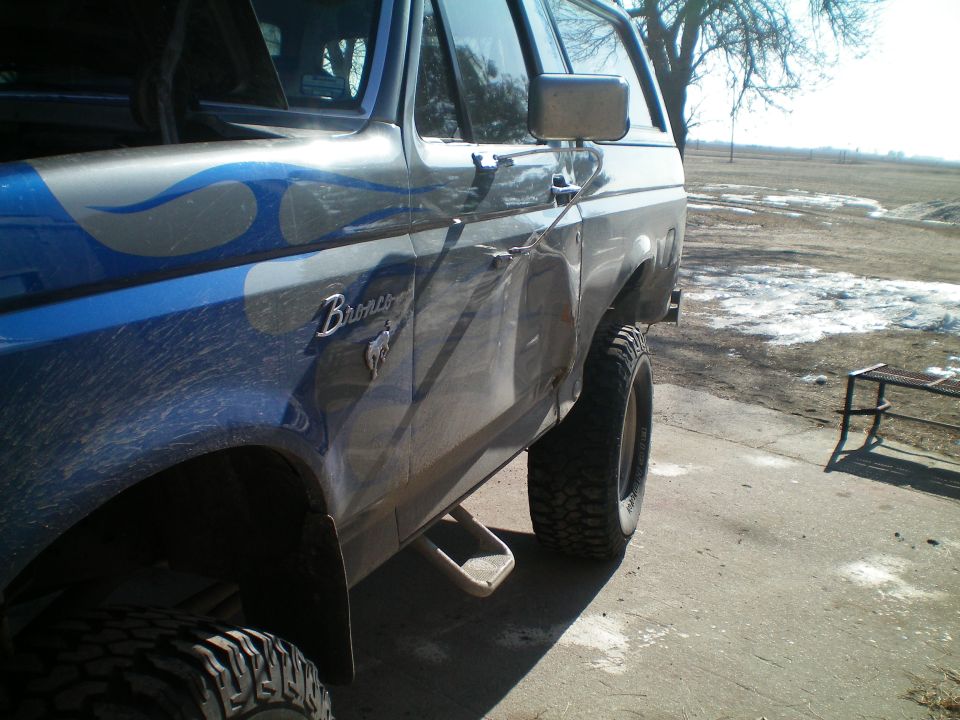 Blue and silver Ford Bronco with flame decals and a dented door, lifted with large tires