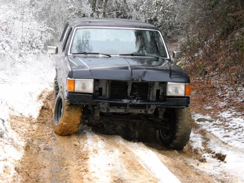 1982 Ford Bronco - This dark blue SUV, likely from the early 1980s, is struggling for traction on a muddy, snow-covered trail.
