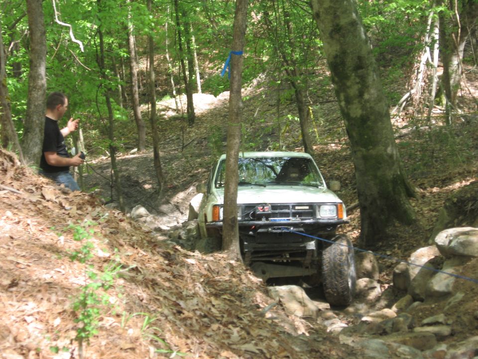 White off-road truck with winch on a muddy, rocky trail in a forest. A person stands to the left