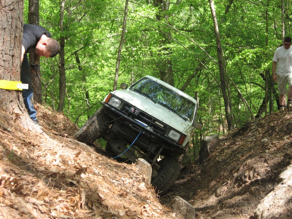 White SUV on a steep, muddy trail with trees and rocks. A person is visible near a tree on the left, and another person is walking on the right
