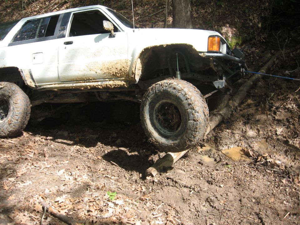 White off-road SUV with large tires stuck in mud and on logs, front right wheel elevated