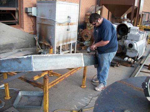 2000 Dodge 1500 - Man uses an angle grinder on a metal beam in an industrial setting with large machinery visible.