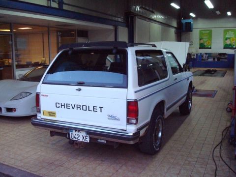 1991 Ford Crown Victoria - A white Chevrolet SUV is parked in a garage next to a white sports car. The SUV has a license plate and 'CHEVROLET' text on the rear.