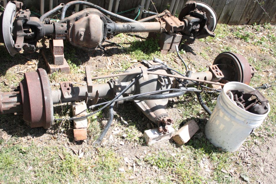 Two vehicle rear axles are sitting on the ground, supported by bricks. A white bucket filled with parts is nearby