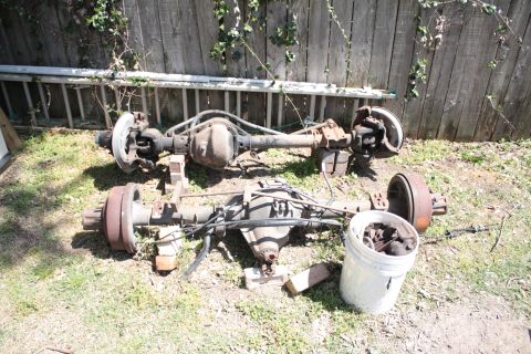 1990 Ford F-150 - Two truck rear axles are shown outdoors, resting on blocks in front of a wooden fence. A bucket of parts is nearby.