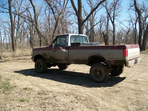 1988 Ford F-150 - This lifted pickup truck, likely from the late 1980s, is covered in mud and parked in a wooded area.
