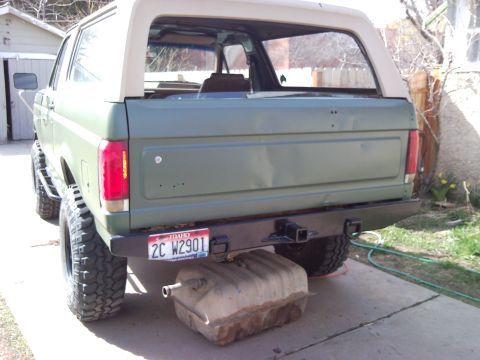 1989 Ford Bronco - Rear view of a green Ford Bronco with a tan top, showing its lifted suspension and a detached fuel tank.