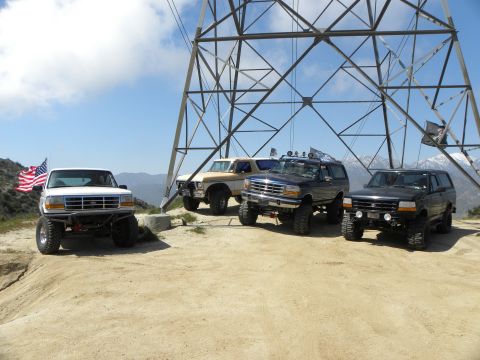 1995 Ford Bronco - Several modified Ford Broncos are gathered on a dusty hillside, with a massive electrical pylon dominating the background.