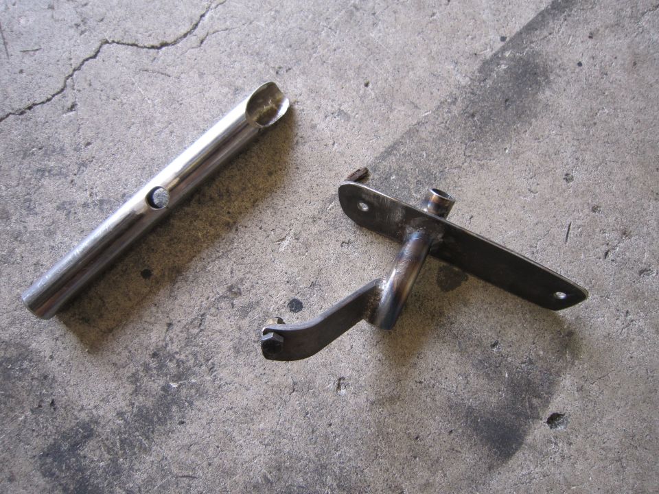 Two metal automotive parts, one cylindrical with a hole, the other a bracket with a lever, lie on a concrete surface