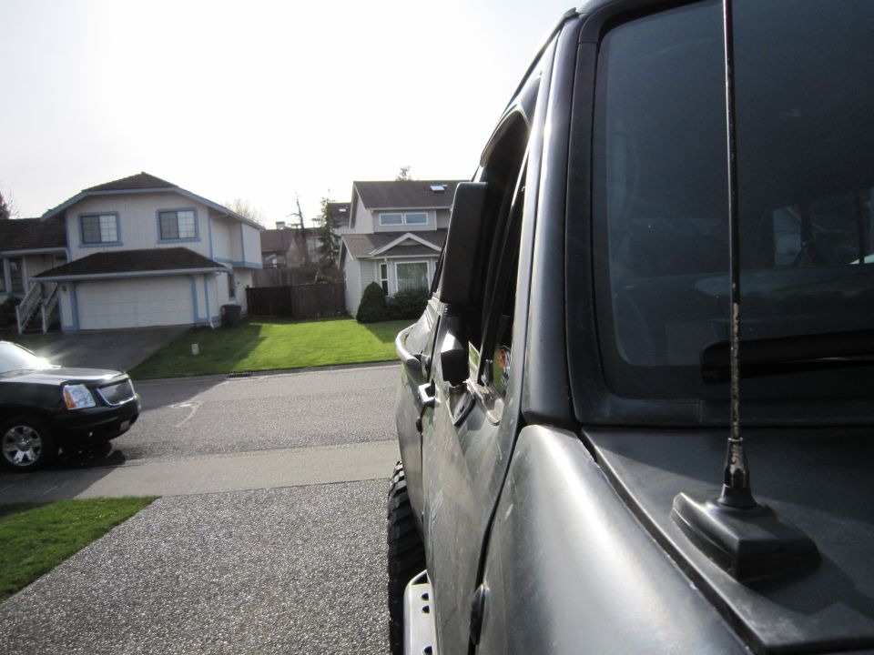 Side mirror and door of a dark SUV parked in a driveway, with another SUV visible across the street