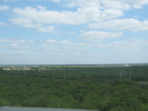 Overhead view of a dense green forest under a blue sky with scattered clouds. A distant structure is visible…