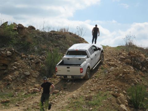 1990 Ford Bronco — White pickup truck with a toolbox in the bed, going up a rocky dirt hill. Guy standing on the roof