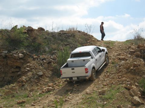 1990 Ford Bronco — White pickup truck with a white cap and silver toolbox driving up a rocky dirt hill with a man standing on th…