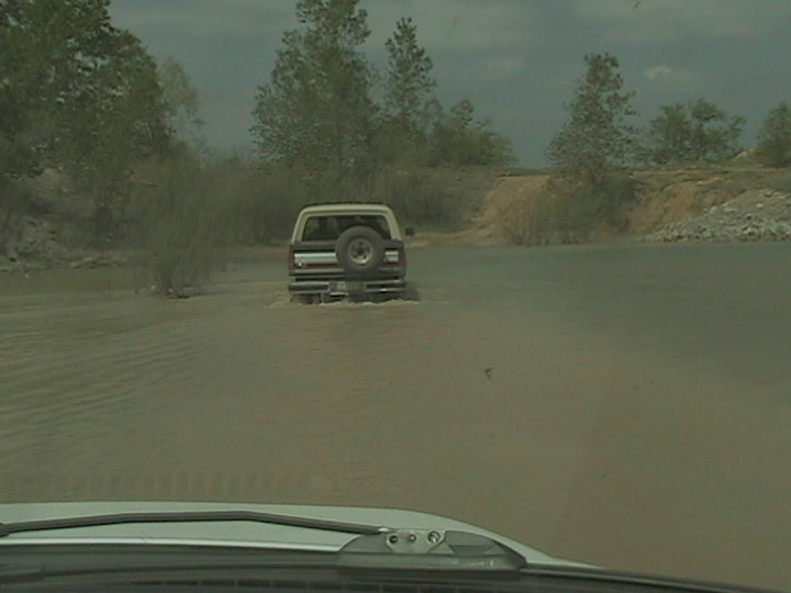 Rear view of a white Ford Bronco driving through shallow water. It has a spare tire mounted on the back