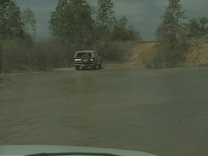 1990 Ford Bronco driving through shallow water on a dirt path