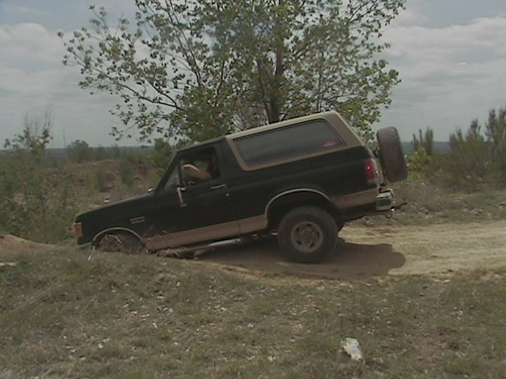 Black Ford Bronco with tan top driving off-road, front driver's side wheel stuck in mud