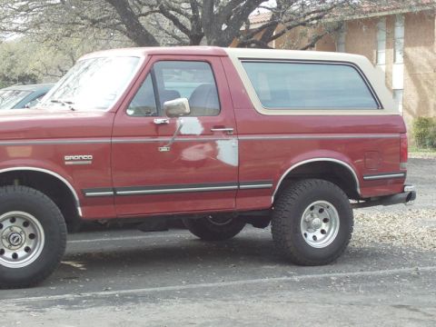 1988 Ford Bronco - Side profile of a red 1988 Ford Bronco a tan removable top and the iconic 'Bronco' emblem on the fender.